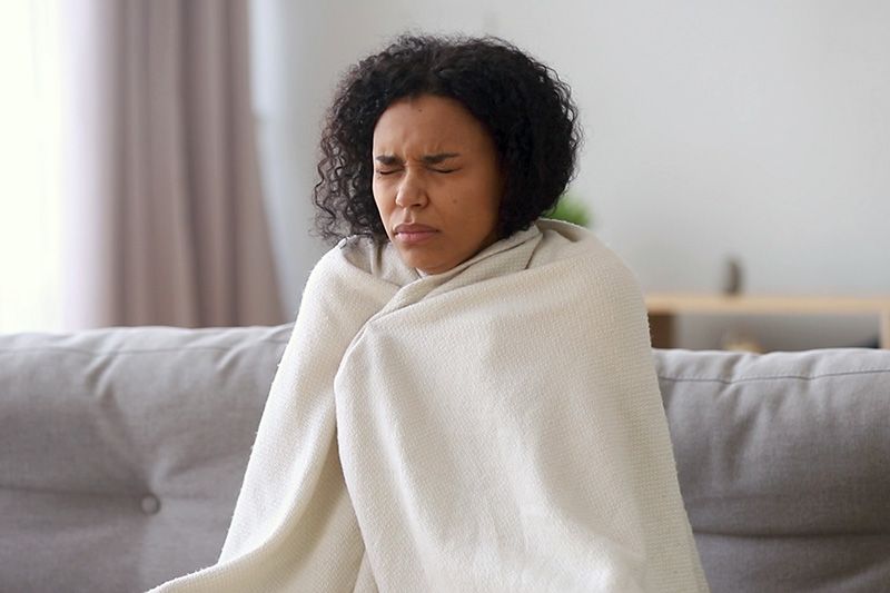 Video - Upgrade Your Furnace. Image shows woman sitting on couch and wrapped in a white blanket looking cold.